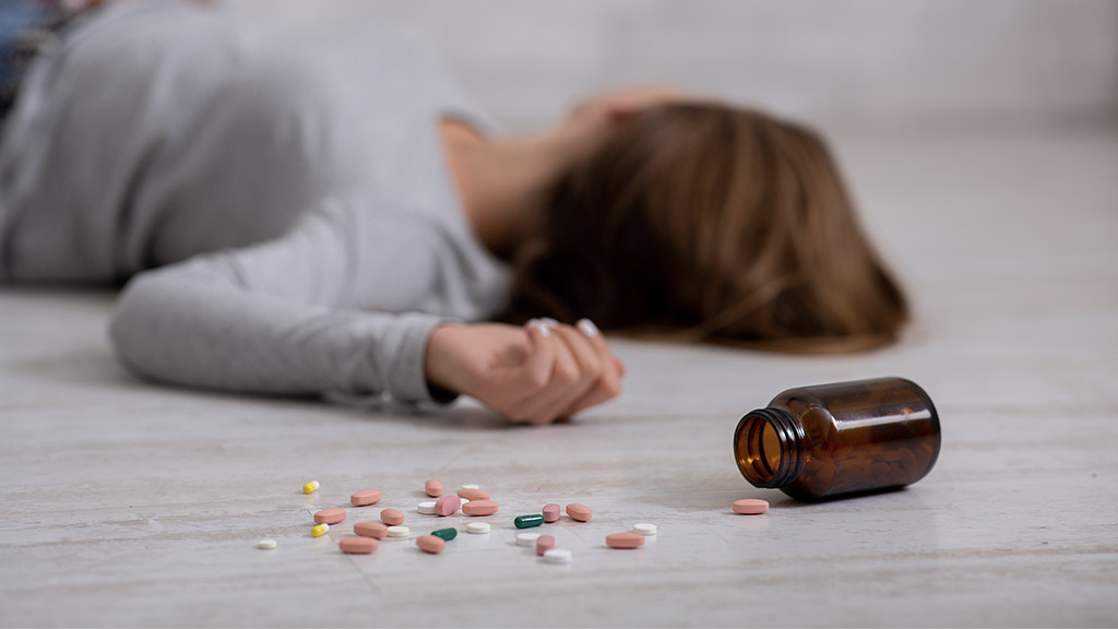 Young woman committing suicide by overdosing on pills, focus on bottle of tablets scattered on floor.