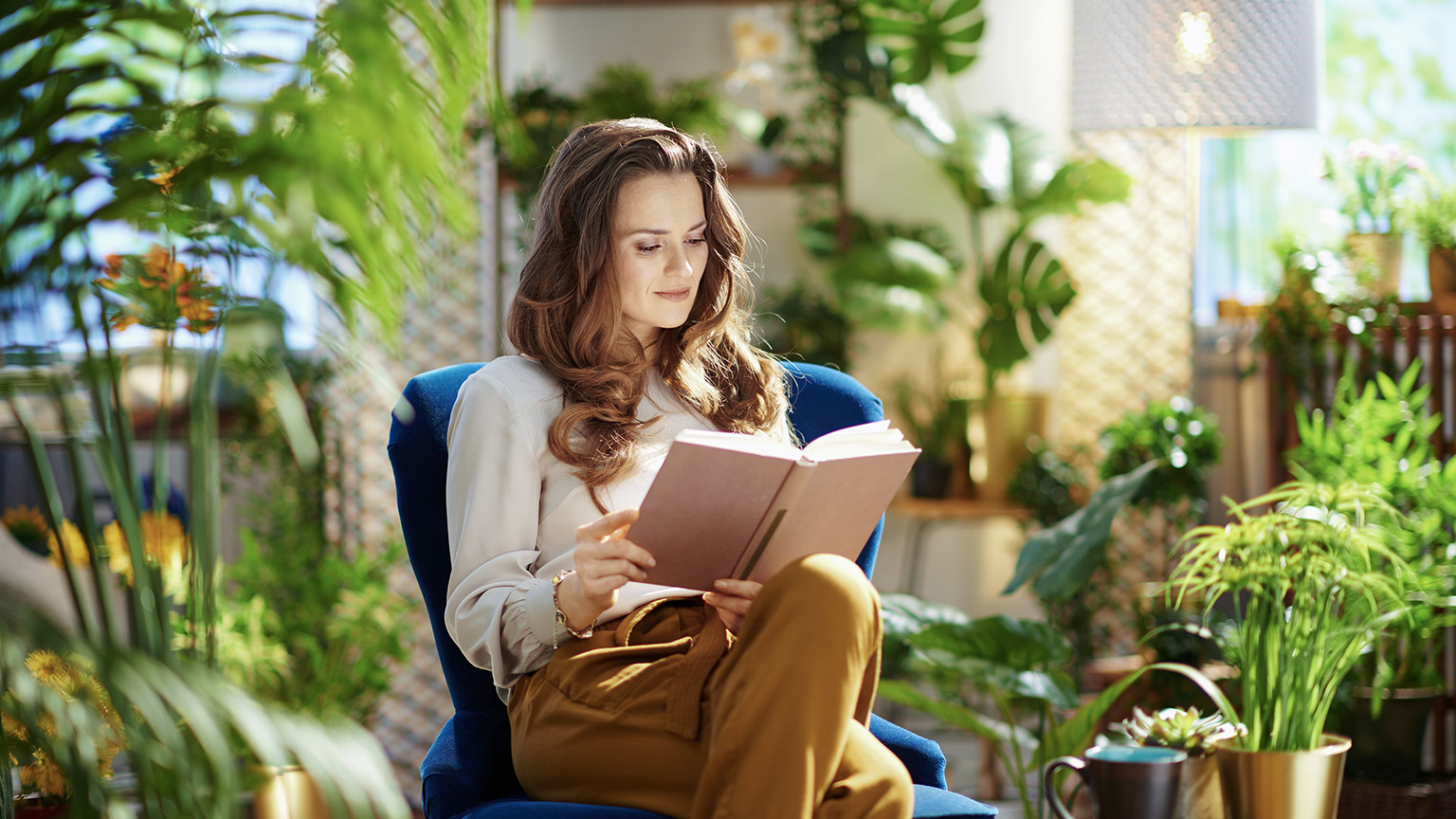 modern female with long wavy hair with book in green pants and grey blouse sitting in a blue armchair in the modern house in sunny day