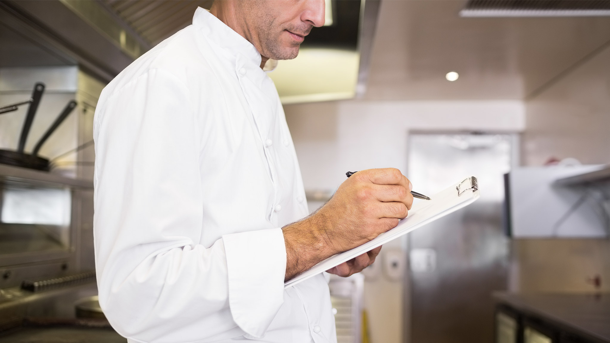 Side view of a concentrated male cook writing on clipboard in the kitchen