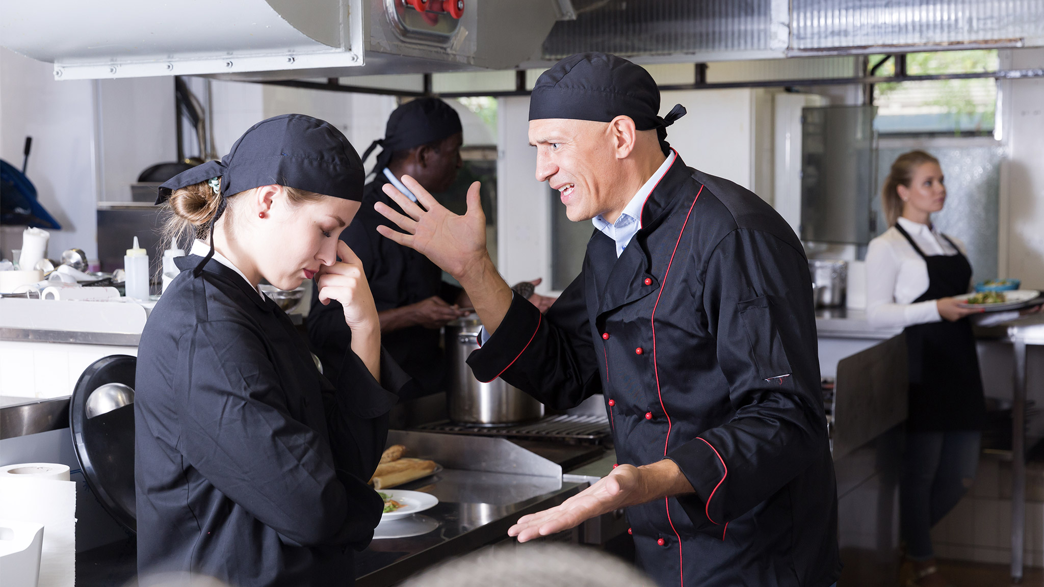 Enraged chef expressing dissatisfaction with work of frustrated girl in restaurant kitchen