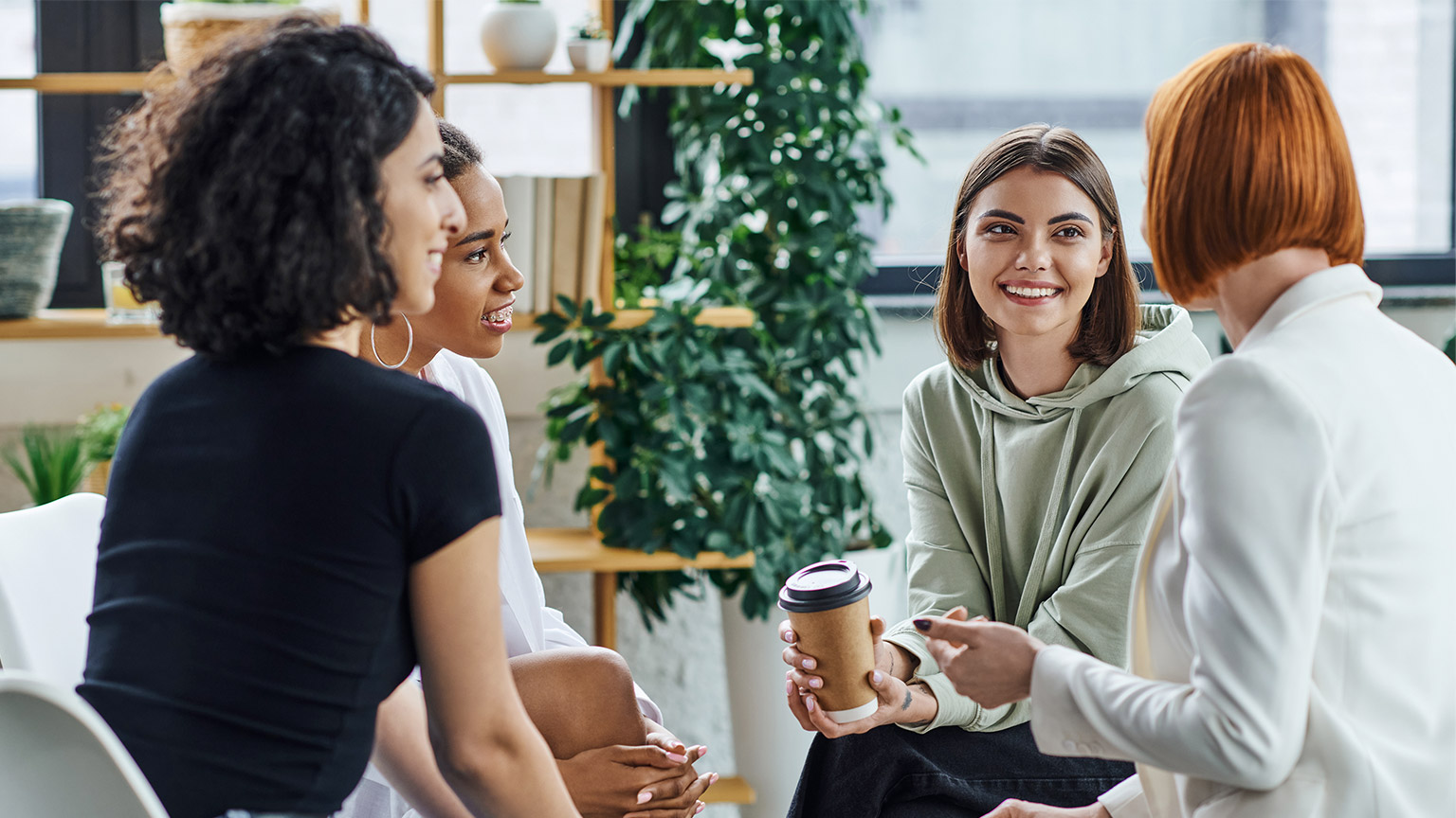 happy young woman sitting with paper cup and listening to redhead psychologist during supportive therapy 