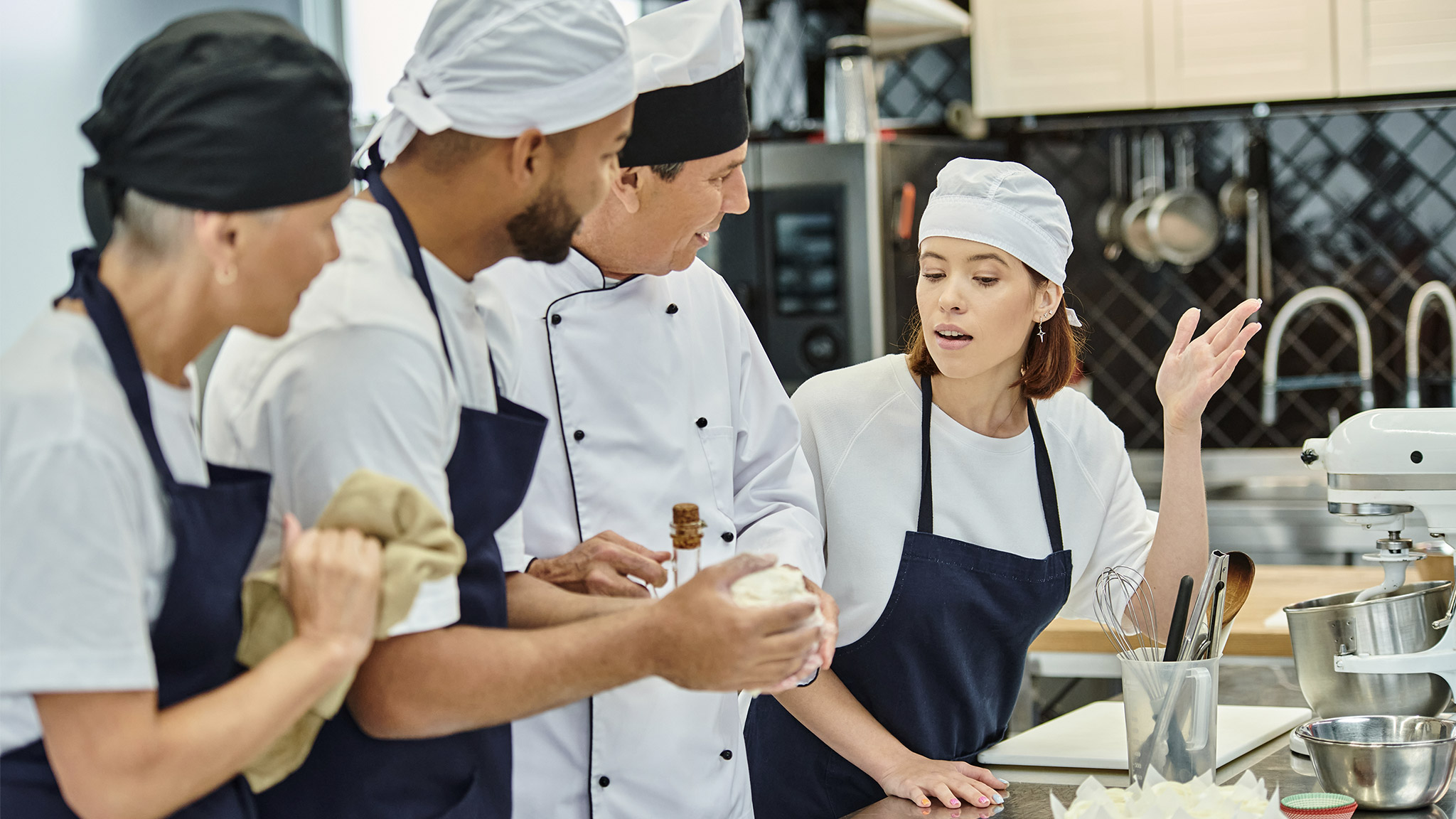 chief cook in white hat smiling at his young chef asking question next to her colleagues