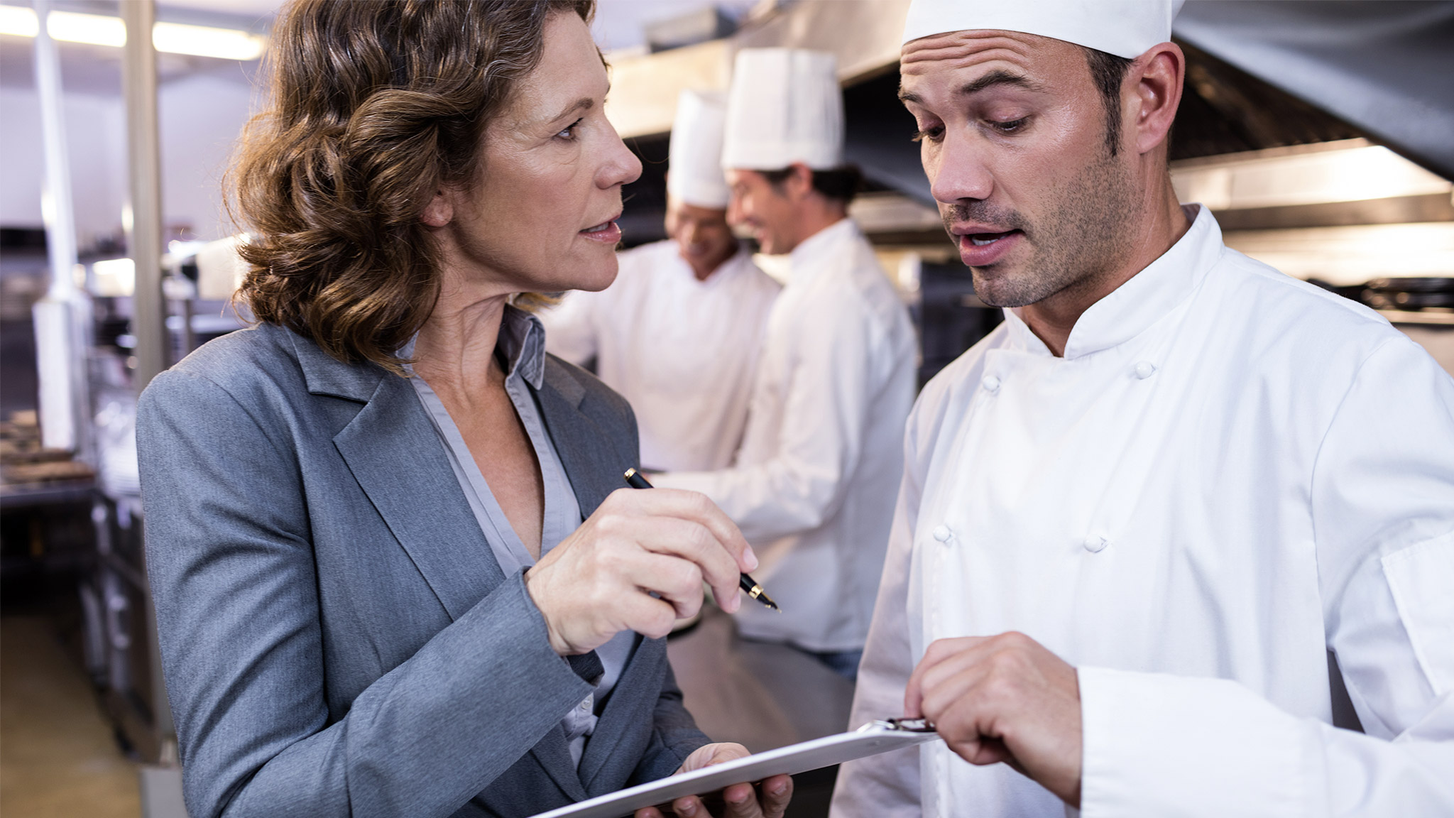  manager writing on clipboard while interacting to head chef in commercial kitchen