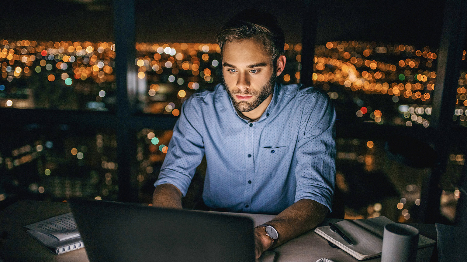 Focused young businessman working on a laptop at his office desk late in the evening in front of windows overlooking the city