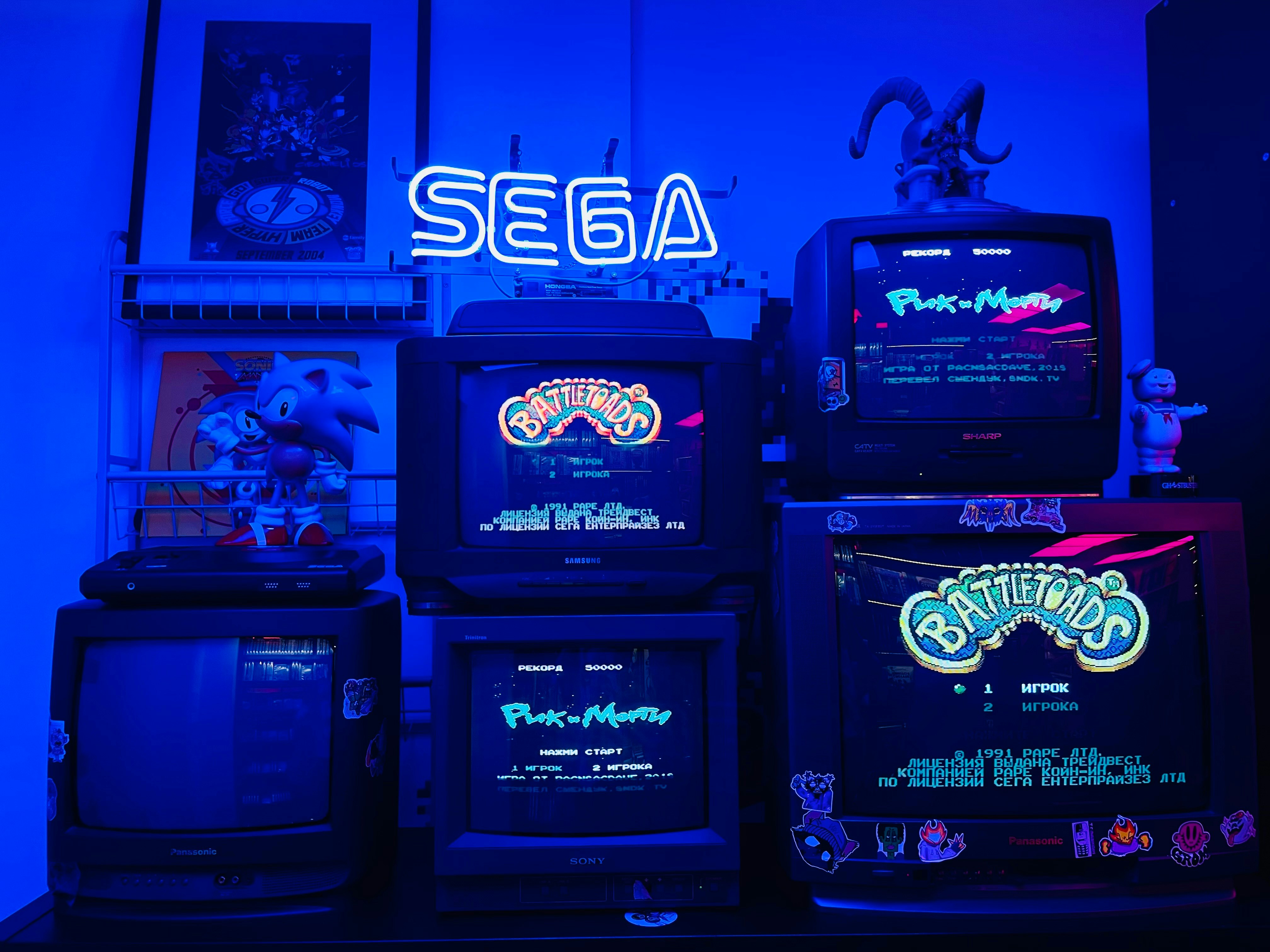 Old CRT televisions are stacked on top of each other, each showing different Nintendo games. The room is bathed in a vibrant blue light from a glowing SEGA sign.