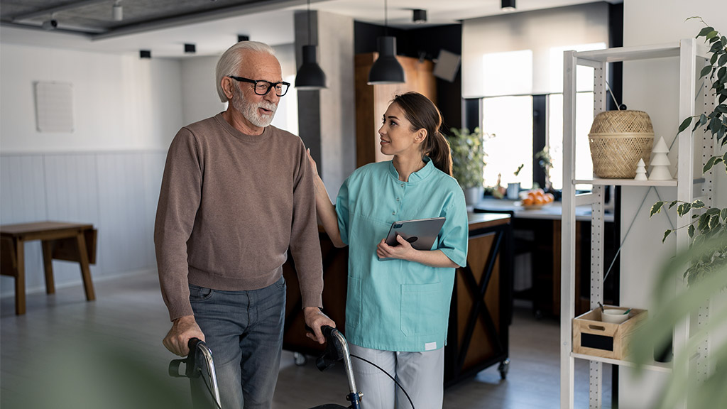 Smiling physiotherapist showing empathy and support to elderly male patient with disability while he's walking with walking frame