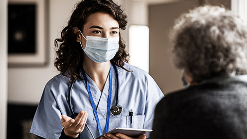 Doctor wearing safety protective mask supporting and cheering up senior patient during home visit during covid-19 pandemic. Nurse and old woman wearing facemasks during coronavirus and flu outbreak. 