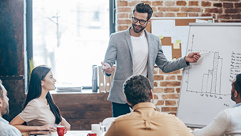 man in glasses standing near whiteboard and pointing on the chart while his coworkersman in glasses standing near whiteboard and pointing on the chart while his coworkers