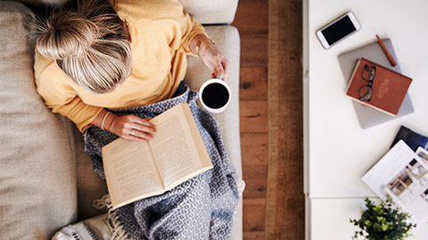 Overhead Shot Looking Down On Woman At Home Lying On Reading Book And Drinking Coffee