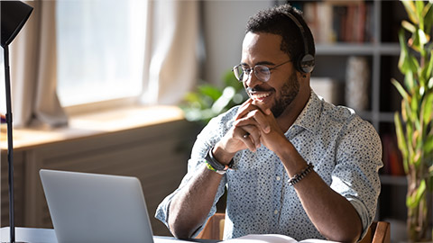 Happy millennial african american man in glasses wearing headphones, enjoying watching educational webinar on laptop