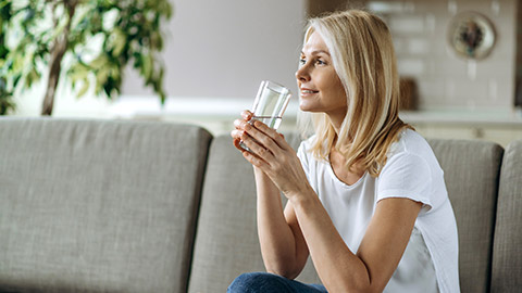 woman sits on the couch in a living room, holding a glass of pure water in a hand