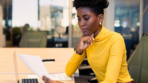 African american businesswoman reading document while sitting with laptop at workplace