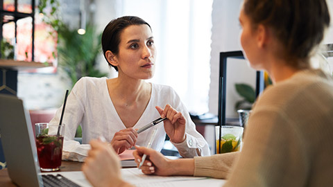 Young serious brunette businesswoman in white blouse looking attentively at colleague while listening to her idea during brainstorm