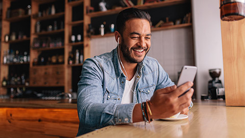 Happy young man sitting at coffee shot with earphones making video call to friend