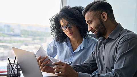 Multiethnic male indian mentor and female African American intern sitting at desk with laptop doing paperwork together