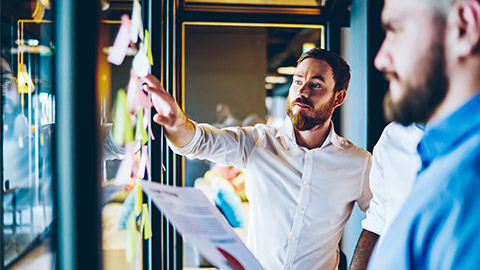 Concentrated bearded interested male employee showing by hand on glass wall with work stickers