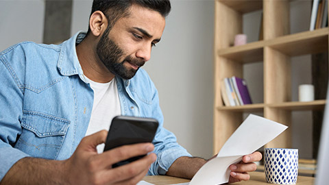 Young indian businessman holding phone reading bank receipt calculating taxes