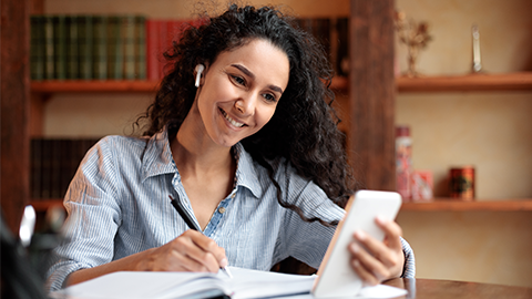 Smiling female student in wireless earphones sitting at desk, using smartphone