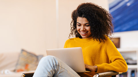 Image of cheerful african american woman using laptop while sitting on chair in living room