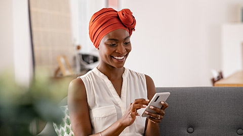 Smiling middle aged african woman with traditional head turban sitting on couch at home using smartphone