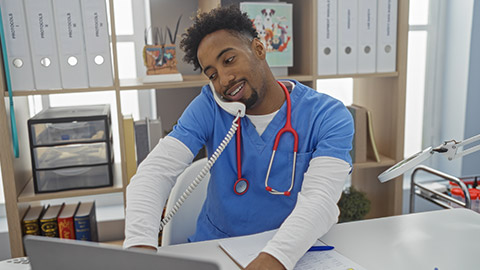 Handsome african american man with a beard working in a veterinary clinic, wearing scrubs and a stethoscope, multitasking with a phone and computer