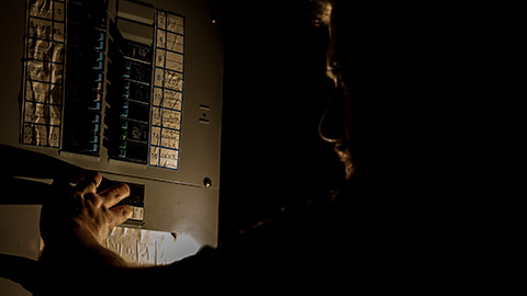 A man, visible only as a faint silhouette, is using a flashlight to investigate a home fuse box in his basement during a power outage.