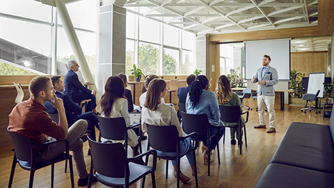 Young berded man speaking on a business training standing in office during a conference or seminar for a group of diverse people company employees sitting on chairs in meeting room