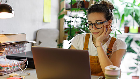 Young female gardener in glasses using laptop, communicates on internet with customer in home