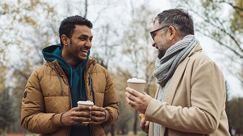 Friends, a senior and a young man walking and talking and drinking coffee together in the autumn park.