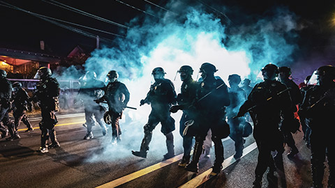 A group of riot police advancing down a street amongst a cloud of tear gas