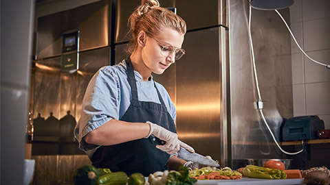 Interested and serious female chef standing in a dark restaurant kitchen next to cutting board while cutting vegetables on it,