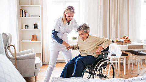 A health visitor helping a senior woman to stand up from a wheelchair.