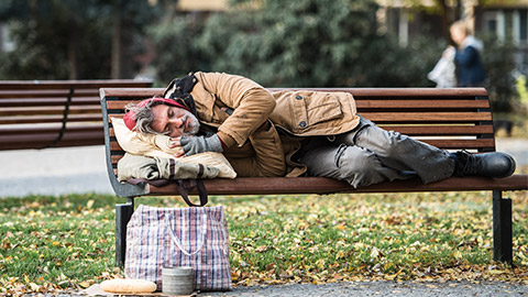 Homeless beggar man with a bag lying on bench outdoors in city, sleeping.