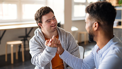 Young happy man with Down syndrome with his mentoring friend celebrating success indoors at school