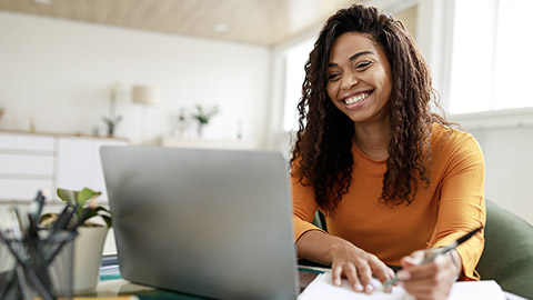 smiling woman sitting at desk, using laptop and writing in notebook, taking notes