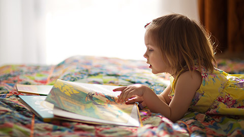 A little cute girl in a yellow dress reading a book lying on the bed