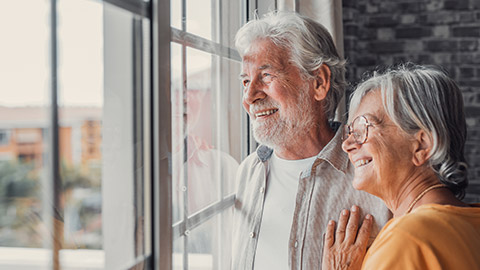 Happy bonding loving middle aged senior retired couple standing near window, looking in distance, recollecting good memories