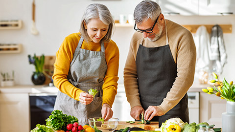 Happy elderly couple smiling husband and wife in aprons prepare salad together at kitchen table