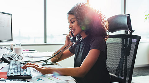 Female business professional sitting at a desk in a modern office working on a computer and talking on the phone