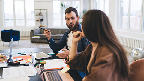 Concentrated young male and female entrepreneurs in casual clothes sitting at table with gadgets and discussing project strategy during meeting