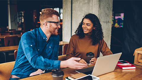 Smiling male teacher with red hair explaining new topic for study to cheerful african american student sitting at laptop computer in coworking