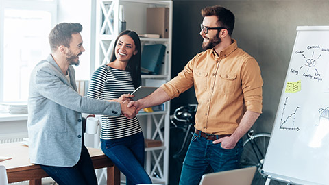 Confident young man standing near whiteboard and shaking hand to his colleague while young woman standing near them and smiling