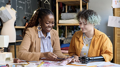 Portrait of young girl with prosthetic arms sewing clothes in inclusive atelier studio with smiling social worker helping