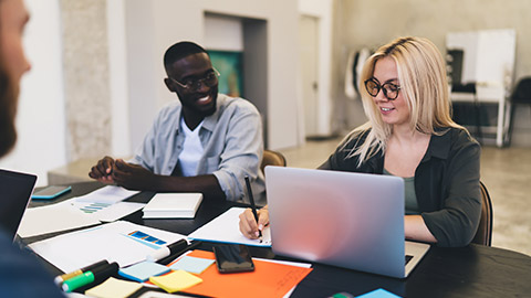 Young multiracial colleagues sitting together at table with laptop and listening to coworker explaining startup strategy during work in office