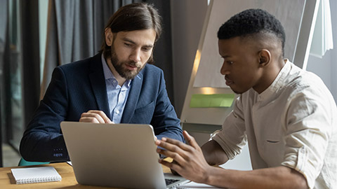 Diverse multiracial male business colleagues sit at desk in boardroom brainstorm negotiate use laptop together, serious multiethnic coworkers talk discuss