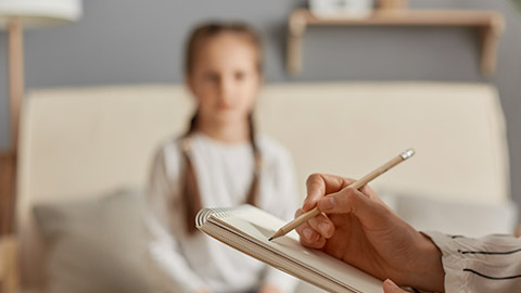 Closeup of doctor psychologist holding paper notebook making notes while talking with small girl at psychological therapy session meeting