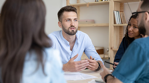 Close up serious bearded businessman telling employees about project sit at table in boardroom at company meeting.