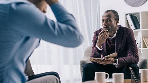 Frustrated young man sititng on sofa and african american psychiatrist in office