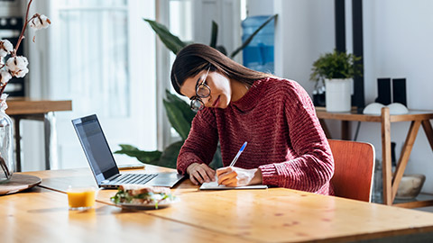 Shot of beautiful young woman working with laptop while writting some notes in the kitchen at home