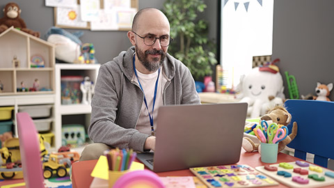 Young bald man preschool teacher using laptop working at kindergarten